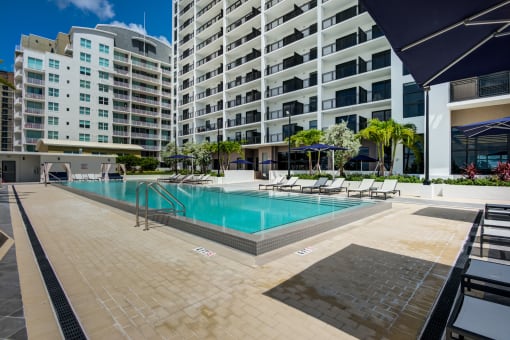 One-Bedroom Apartments in Miami, FL - MB Station - Sparkling Swimming Pool Surrounded by Lounge Seating, Palm Trees, Raised Planters, and an Apartment Building in the Background.