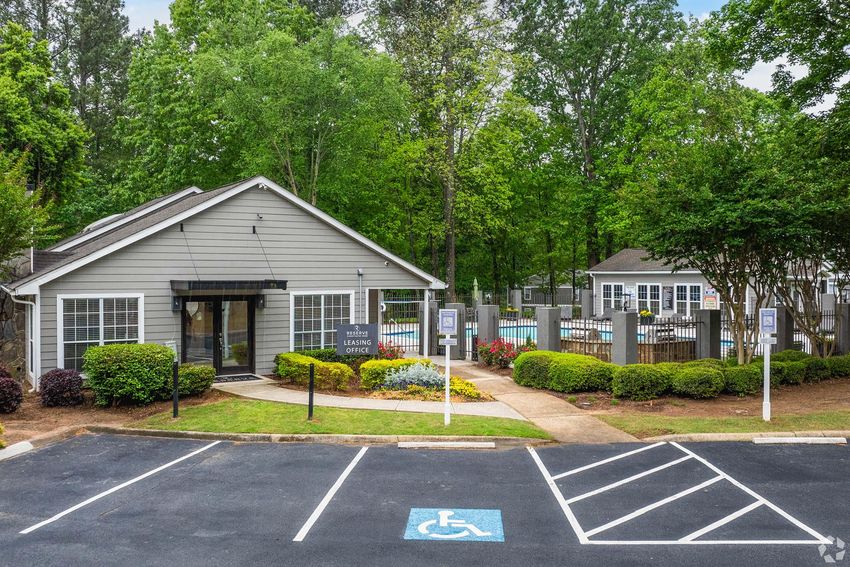 A view of an apartment complex's leasing office surrounded by landscaped gardens and trees. In the foreground is a parking lot with a wheelchair-accessible space marked. Visible amenities include a swimming pool in the background with a fenced area and signposts indicating various facilities.