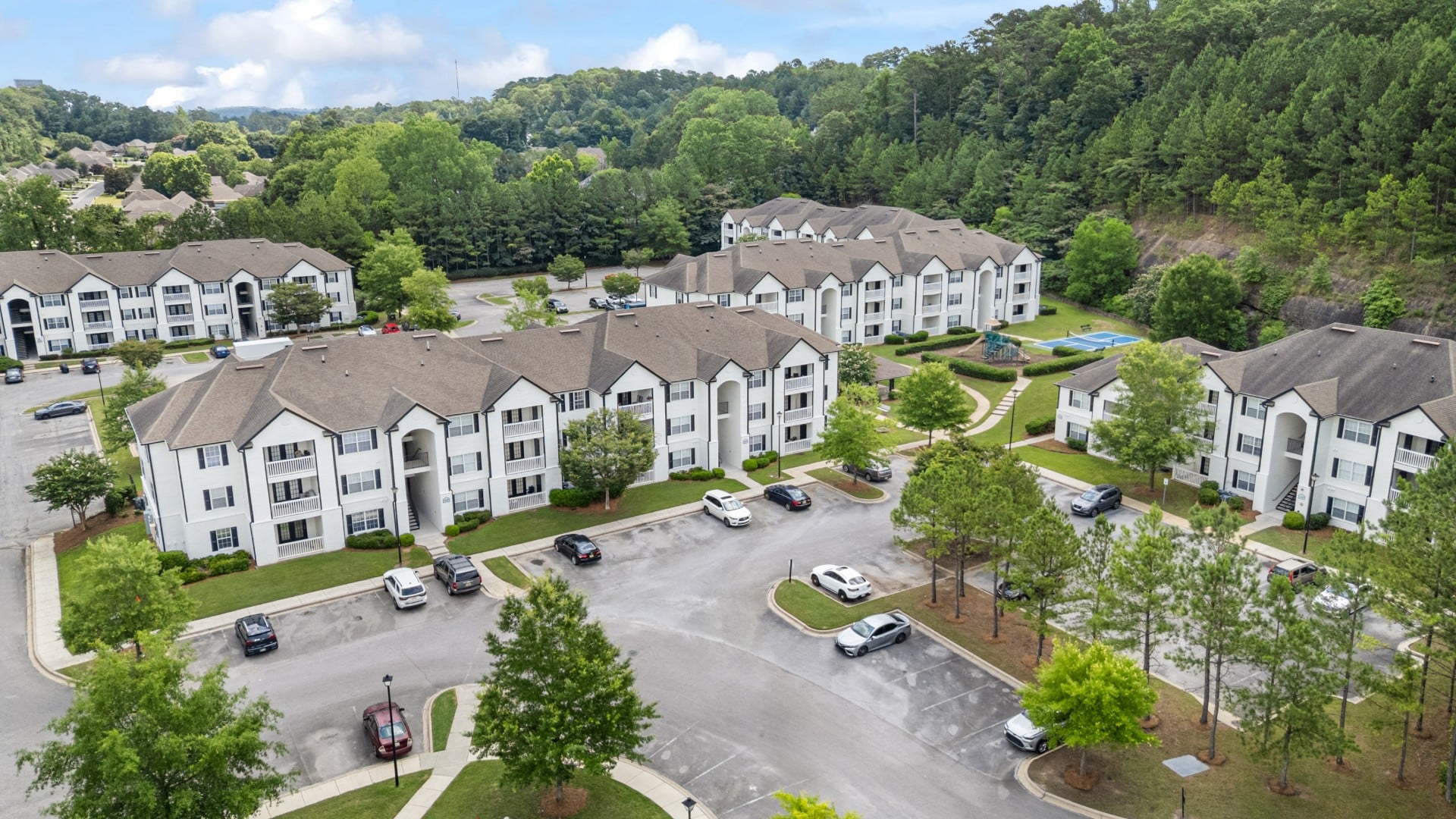 Aerial view of a residential apartment complex with multiple white buildings, parking lots, green lawns, and trees surrounding the area.