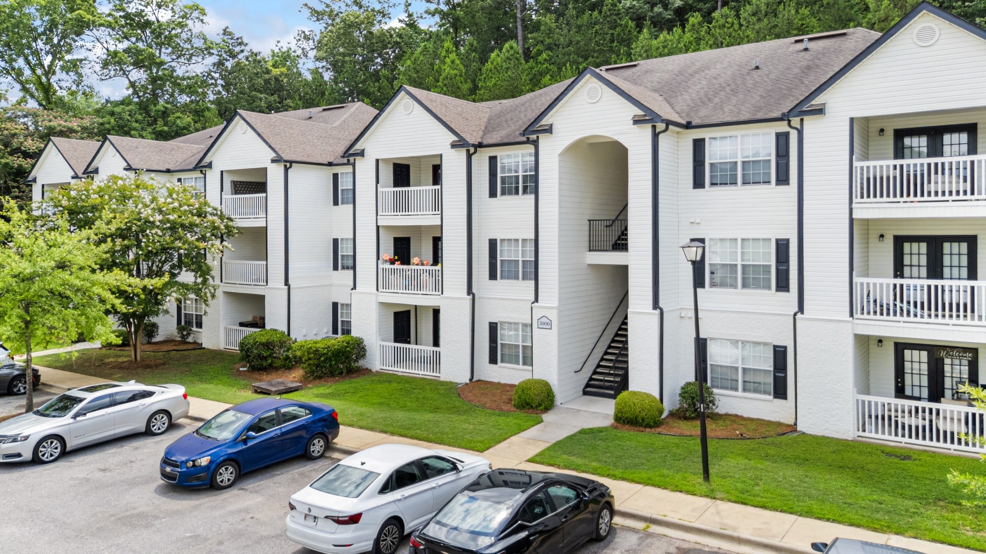 Three-story white apartment building with balconies and exterior staircases, surrounded by trees, with several parked cars in front on a paved lot.