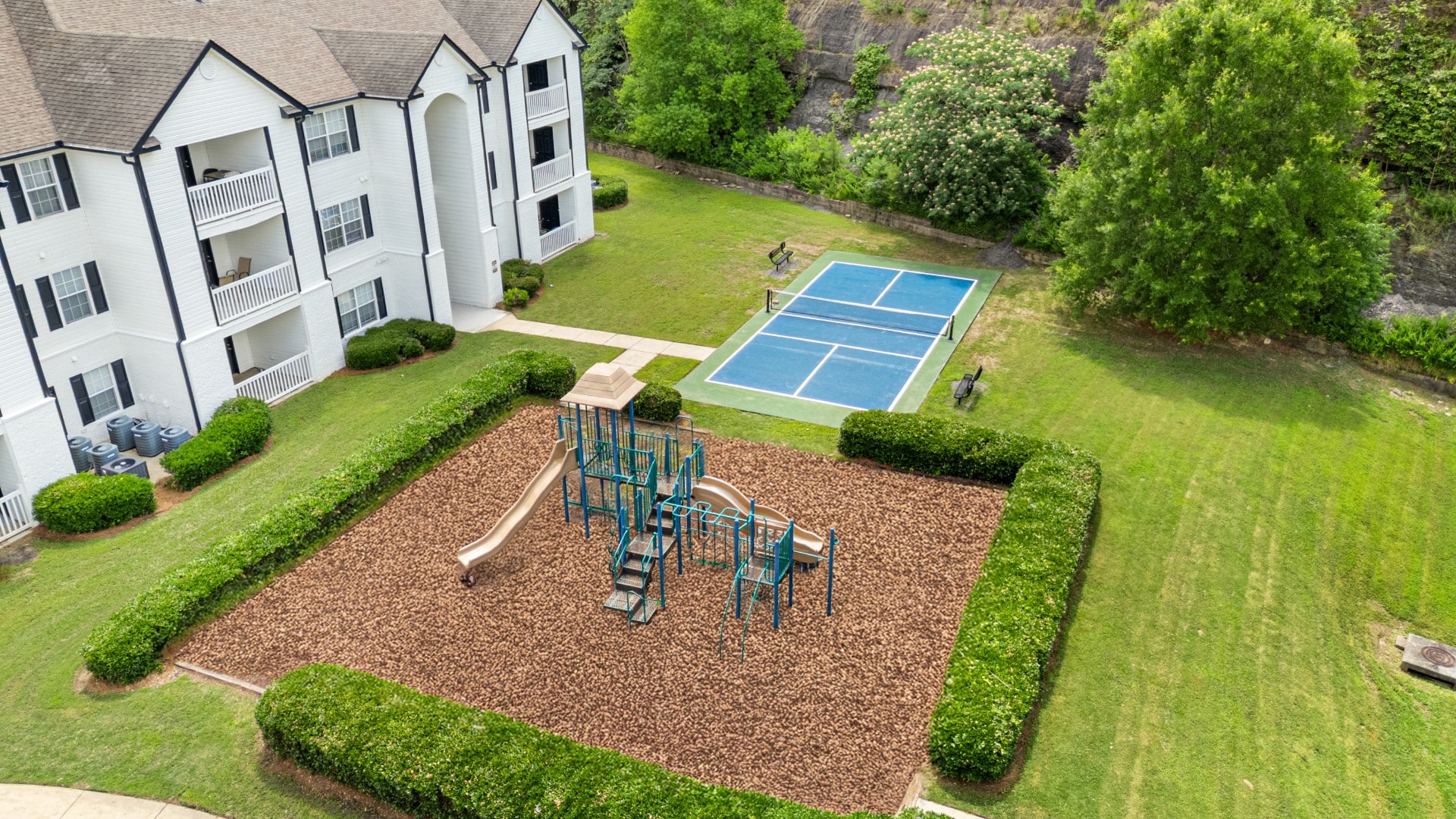 Aerial view of The Ridgely residential complex with a playground, a blue pickleball court, and green lawns surrounded by trees and shrubs.