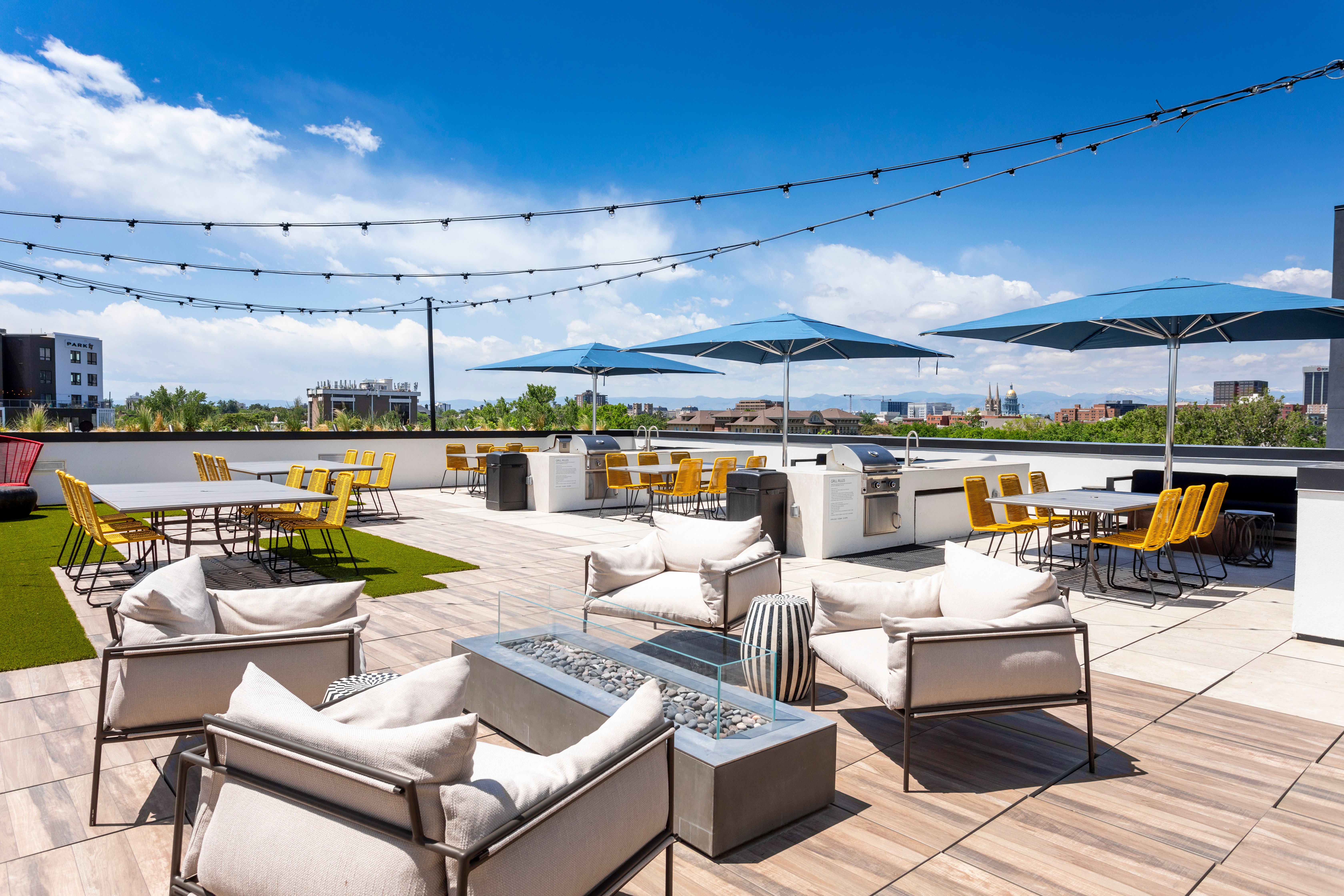 A modern rooftop terrace with seating areas, an outdoor fireplace, and blue umbrellas. The sky is clear and sunny, creating a bright atmosphere.