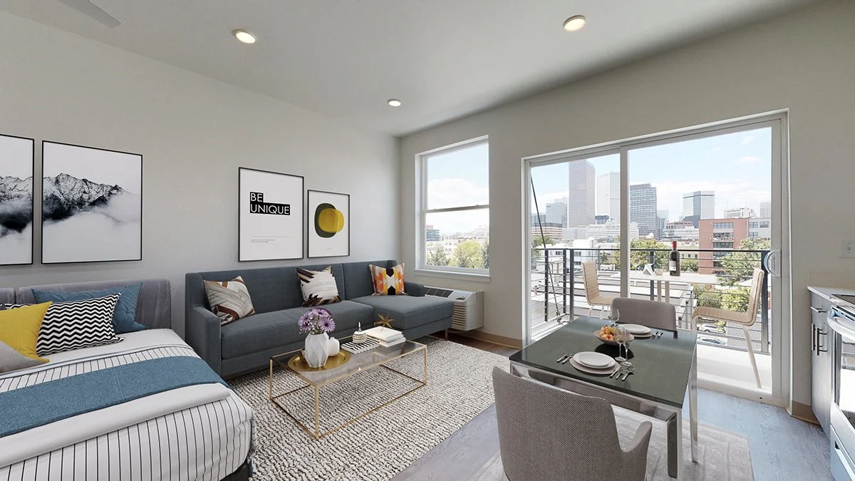 A modern studio apartment showcasing a cozy living area with a gray sofa, a stylish coffee table, and a bed with striped bedding. Large windows allow natural light to flood the space, offering a view of the city. A dining area is set for two with a small table and chairs, complemented by contemporary art on the walls.