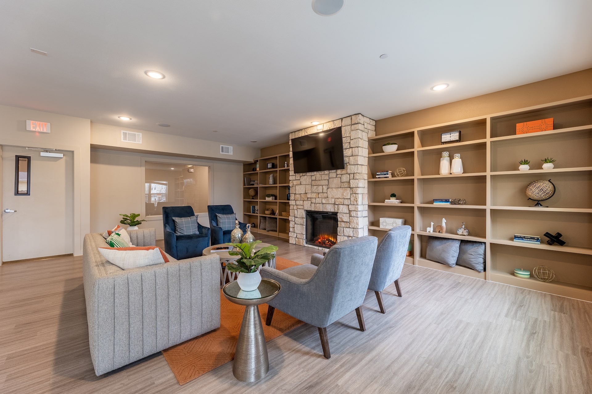 A resident lounge with four blue chairs and a beige couch, next to a fireplace and a TV.