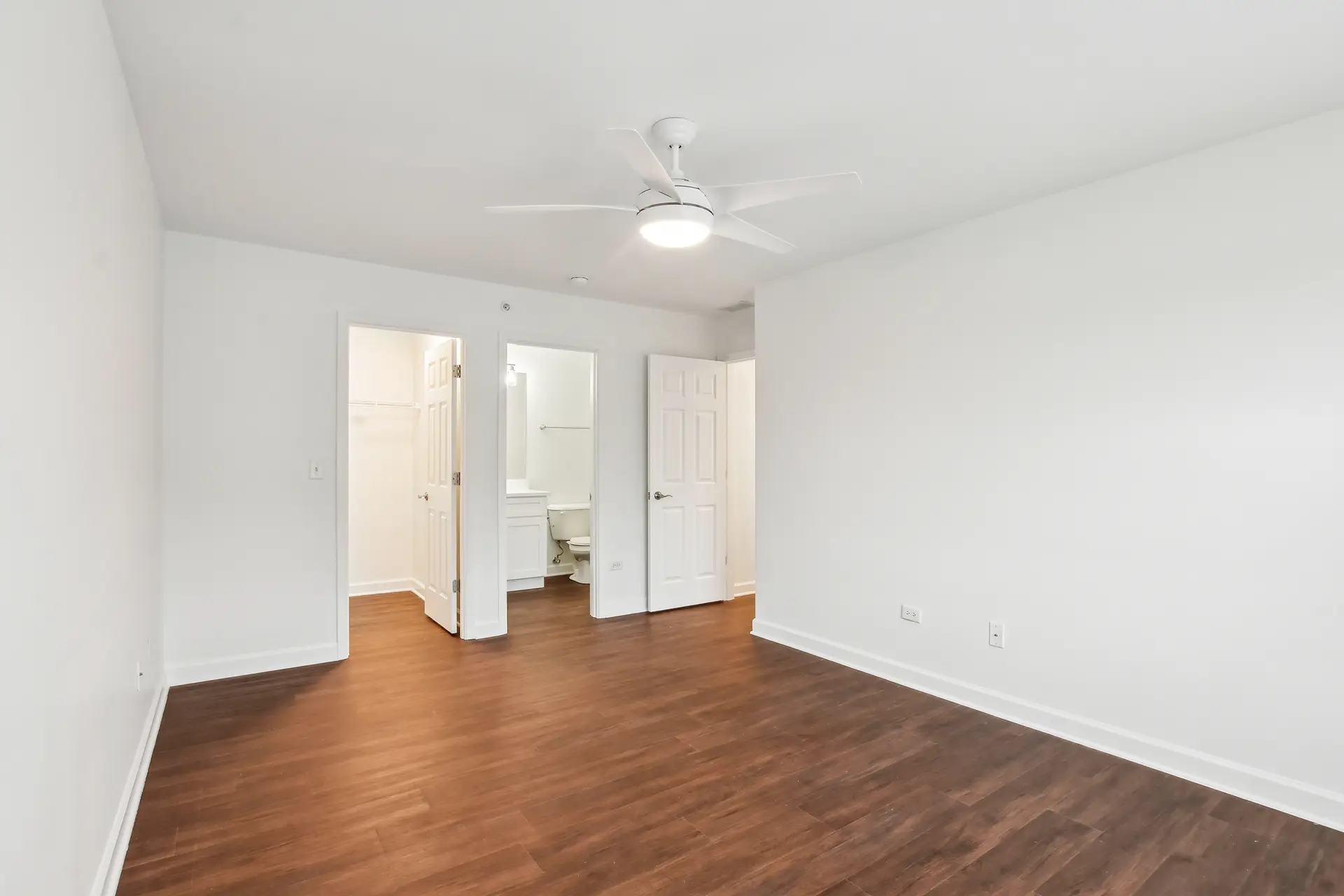 Empty walk-in closet with white walls, white wire shelves on two sides, and dark wood-style flooring. Door partly open to a clean space.