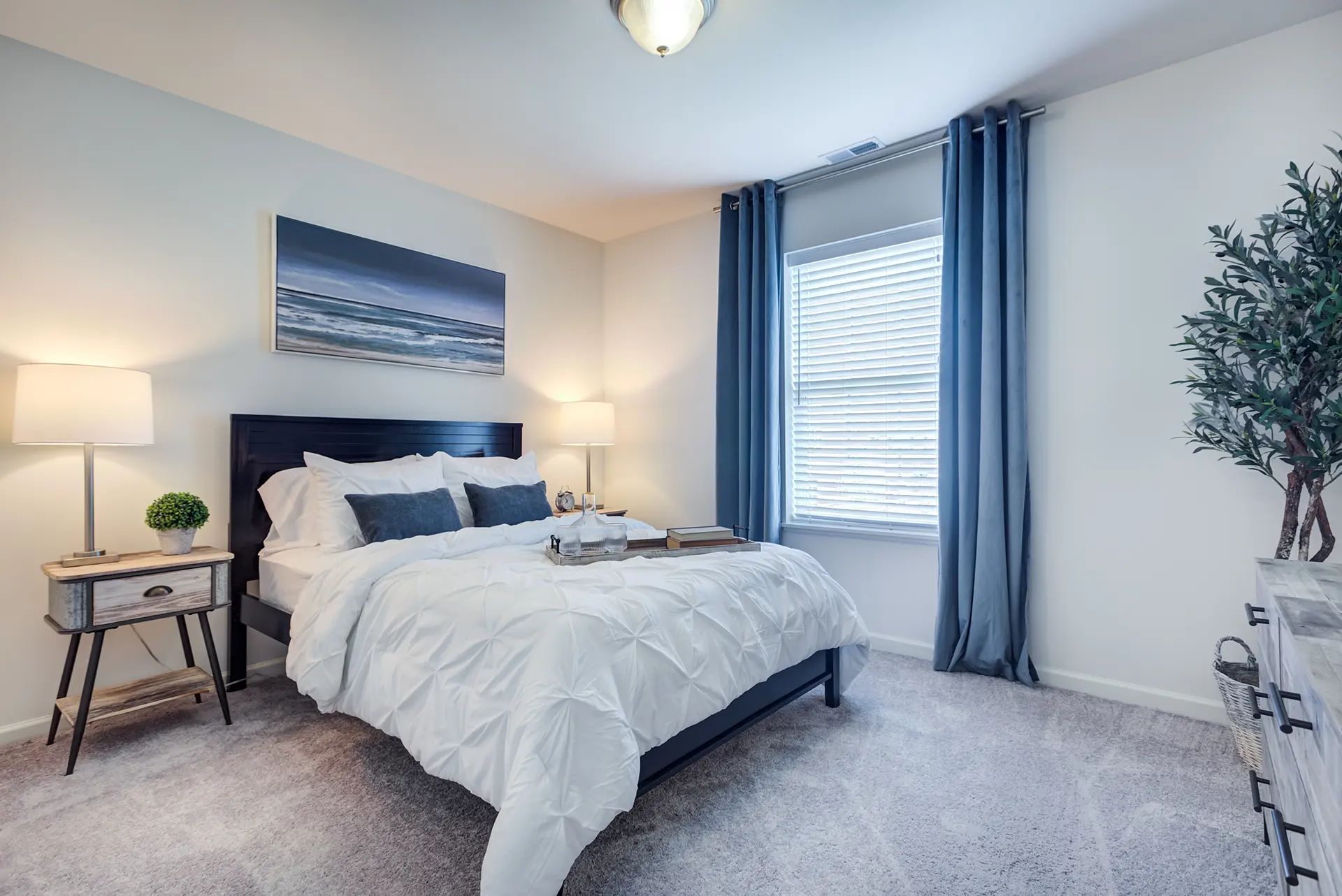 A modern bedroom with a neatly made bed, white bedding, gray and blue pillows, two wood-like bedside tables with lamps, a large window with blue curtains, a beach-themed painting, and a potted plant in the corner.