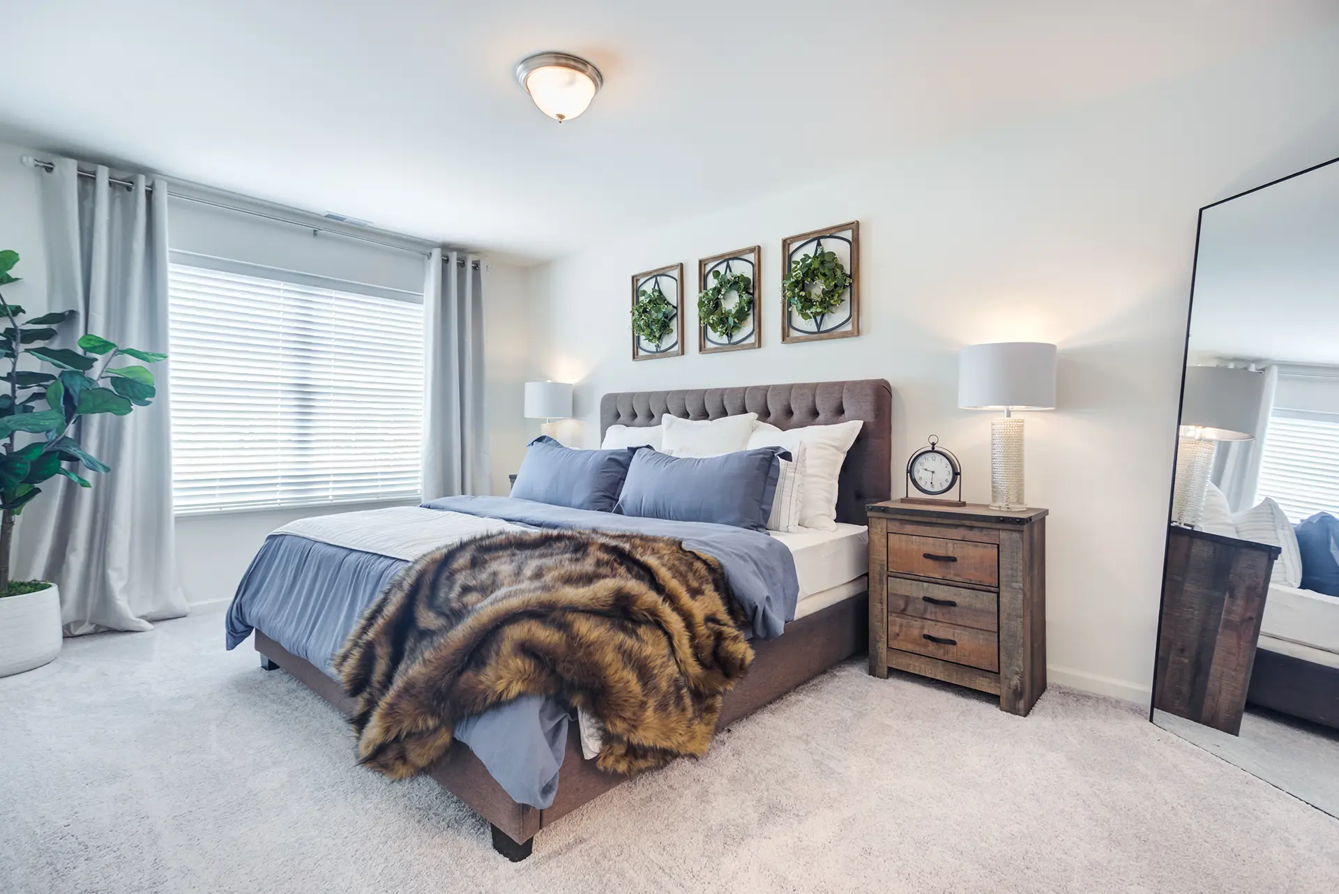 A modern bedroom with a large bed, blue and white bedding, faux fur throw, wood-look bedside tables with lamps, plant, mirror, art. Light from a window with gray curtains.