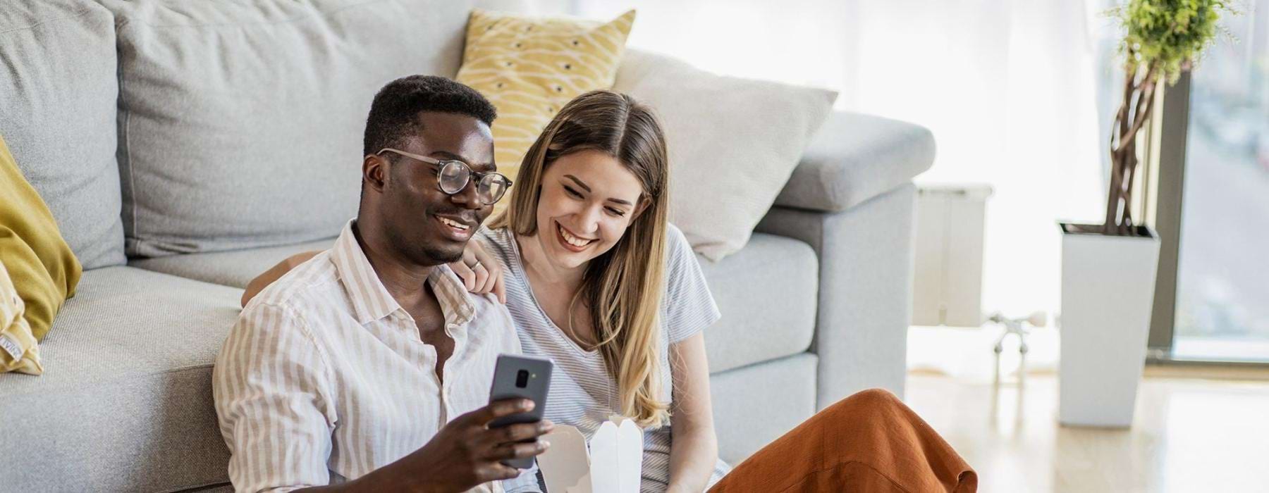 a man and woman with take out, sit against a couch on their living room floor and watch at their cell phone