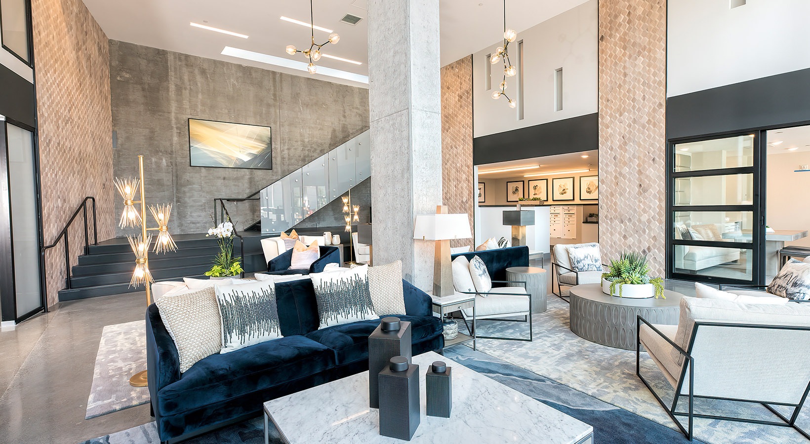 Apartments in Oakland, CA, for Rent- Orion - Lobby with Concierge Featuring Modern Elements Such as a Velvet Sofa with Pillows, a Coffee Table, an Area Rug, and Multi-Bulb Pendant Lights.