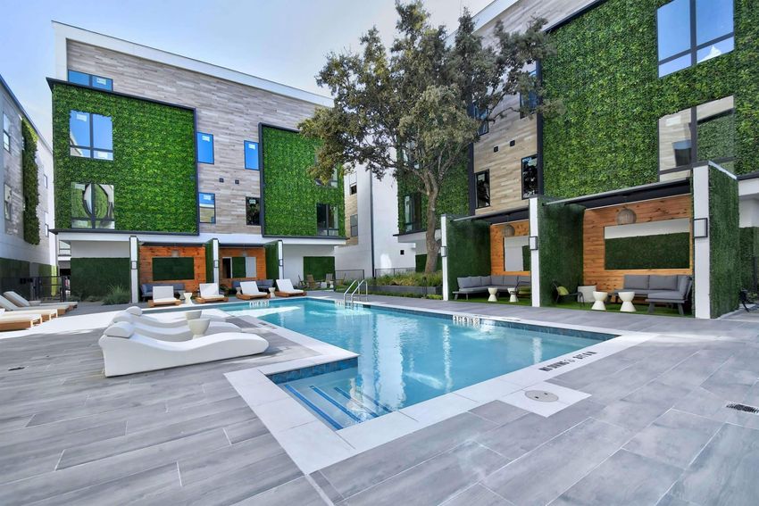 Modern outdoor pool area surrounded by sleek, contemporary buildings with green foliage accents. The pool features lounge chairs and shaded cabanas, creating a stylish and inviting atmosphere for relaxation. Bright sunlight illuminates the space, enhancing the vibrant greenery and elegant design.