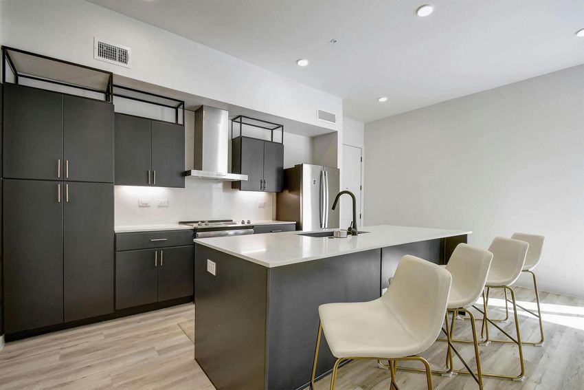 Modern kitchen featuring sleek black cabinetry, stainless steel appliances, and a large island with a white countertop. Four white bar stools are positioned at the island. The space has bright, neutral walls and recessed lighting, creating a clean and contemporary atmosphere.