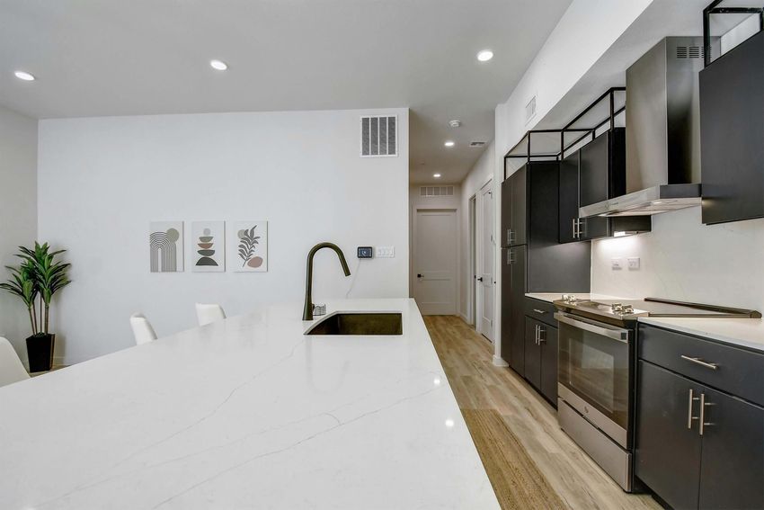 Modern kitchen featuring a marble island with a sink, sleek black cabinetry, and stainless steel appliances. To the left, there are three decorative wall art pieces. The kitchen is well-lit with recessed lighting and has a minimalist, contemporary design. A plant adds a touch of greenery to the space.