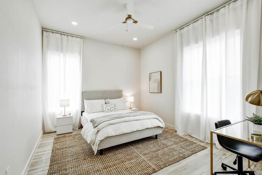 A modern, bright bedroom featuring a queen-sized bed with light bedding, two bedside lamps, and large windows with sheer curtains. There's a small desk and chair in the corner, complemented by a textured area rug on the wooden floor, creating a cozy and stylish atmosphere.