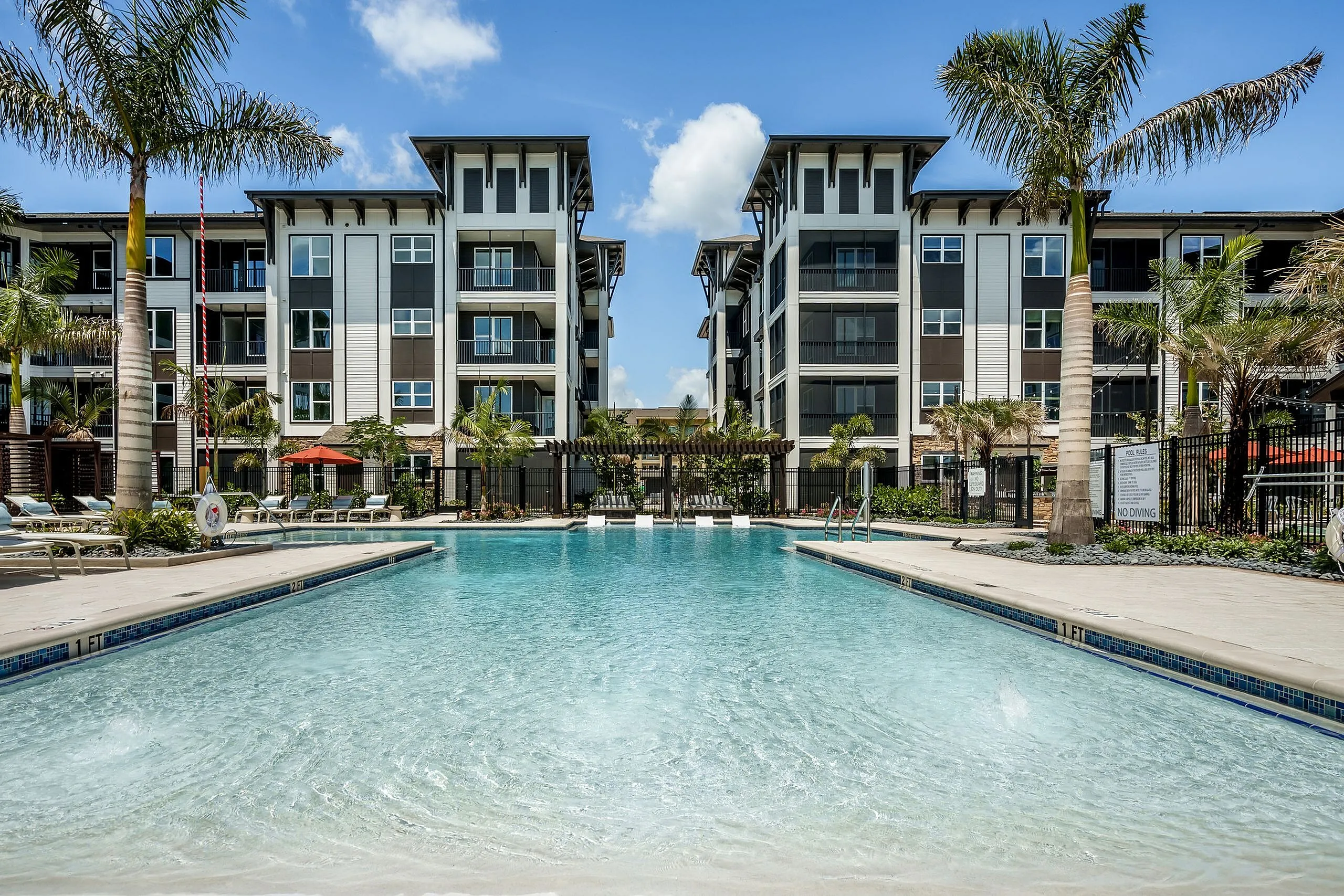 Luxurious apartment complex with modern architecture, palm trees, and a sparkling swimming pool under a clear blue sky.