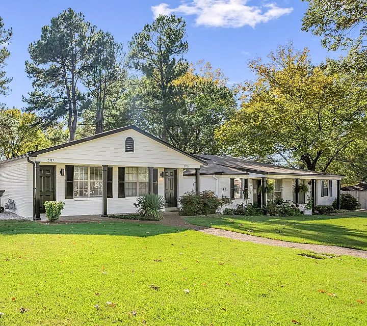 Charming duplex with white brick exterior and lush green lawn, surrounded by mature trees in a serene neighborhood.