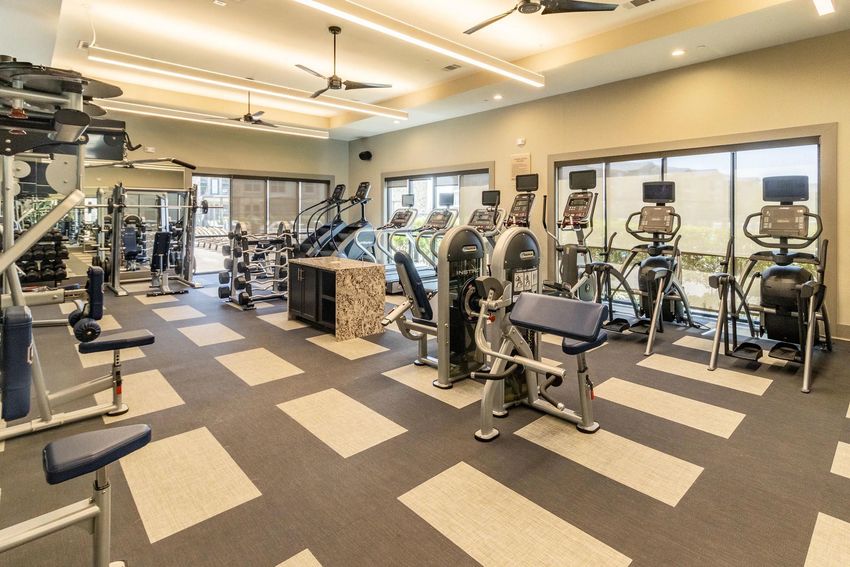 A modern gym space featuring various exercise equipment, including treadmills, elliptical machines, weight machines, and free weights. The room is well-lit with ceiling fans and large windows, providing a view of the outdoors. The flooring is a combination of carpet and tile, enhancing the fitness environment.