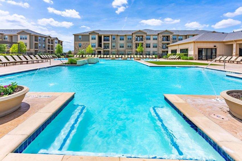 A clear, blue swimming pool surrounded by lounge chairs, with a landscaped area and apartment buildings in the background under a bright blue sky with fluffy clouds. Water features are visible at the pool's edge, adding to the inviting atmosphere of the outdoor space.