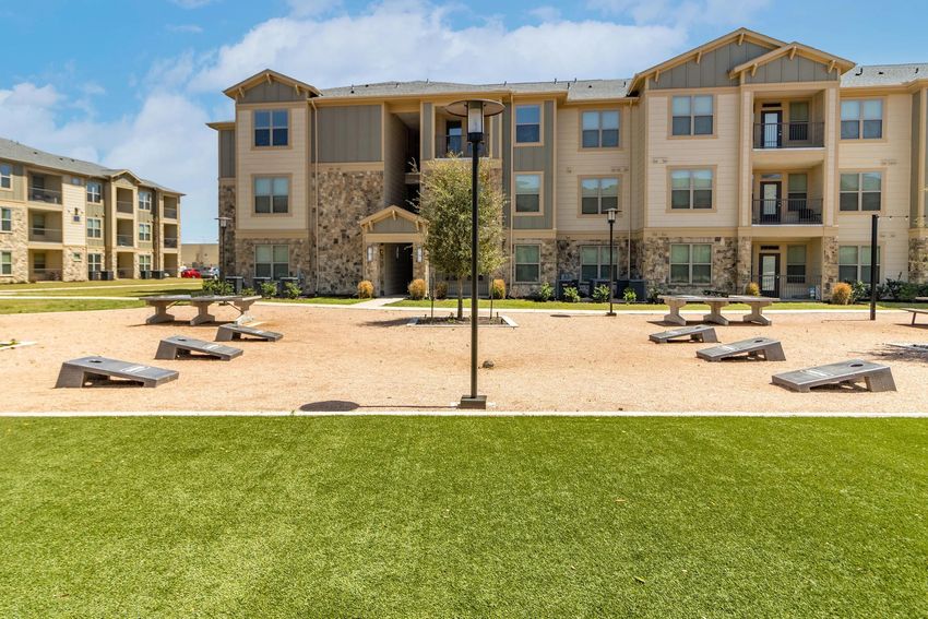 A landscaped courtyard with artificial grass and gravel pathways. In the center, there are several game tables for activities. Surrounding the courtyard are modern apartment buildings with a mix of stone and siding exteriors. The sky is clear with a few clouds.