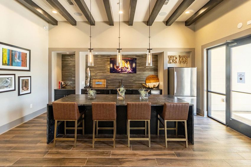 Modern kitchen interior featuring a large dark stone island with four brown leather bar stools. The space includes wooden beams on the ceiling, stylish pendant lights, and decorative artwork on the walls. A sleek modern fireplace is visible in the background, creating a cozy atmosphere. Large windows allow natural light in.