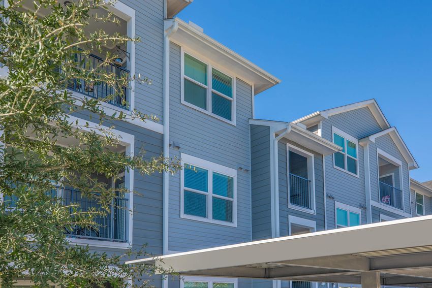 Modern multi-story apartment building with gray siding, featuring several windows and balconies. A green tree is partially visible in the foreground, and a shaded carport structure is located at the bottom of the image. The sky is clear and blue.