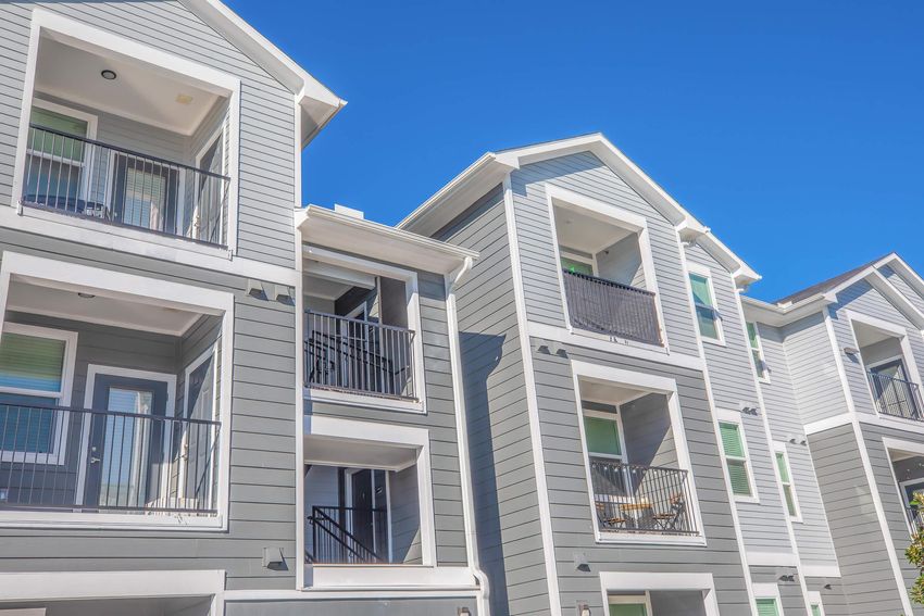 Modern apartment buildings with multiple levels, featuring grey siding, balconies, and large windows under a clear blue sky.