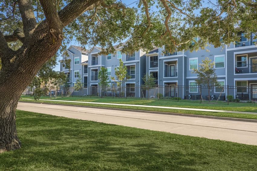 Modern apartment building with multiple units, featuring large balconies and green landscaping. A tree is visible in the foreground, providing shade, with a pathway and manicured lawn along the side. The setting is bright and sunny, suggesting a well-maintained residential area.