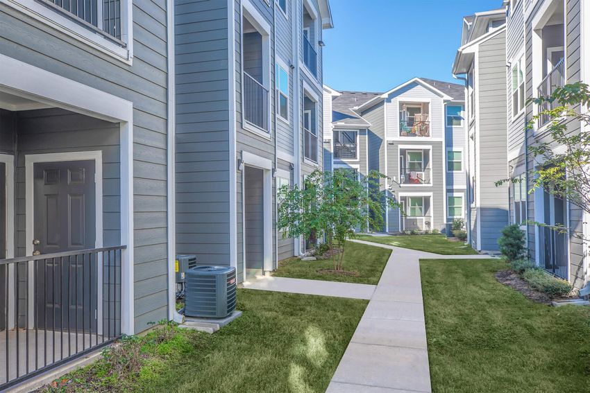A view of a modern apartment community with multiple buildings. The scene shows a well-maintained pathway lined with green grass and shrubs, leading between gray and blue exterior structures. Balconies are visible on upper floors, and the sky is clear and sunny, creating a welcoming atmosphere.