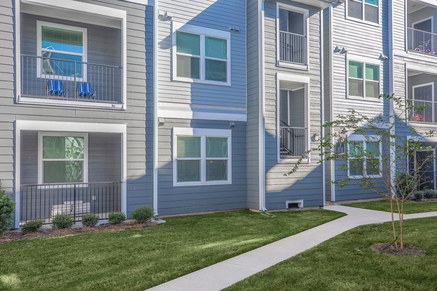A modern apartment community featuring multiple gray and white buildings. The exterior includes balconies with chairs, neatly landscaped grass, small shrubs, and a paved walkway leading through the area. The setting is bright and well-maintained, suggesting a comfortable living environment.