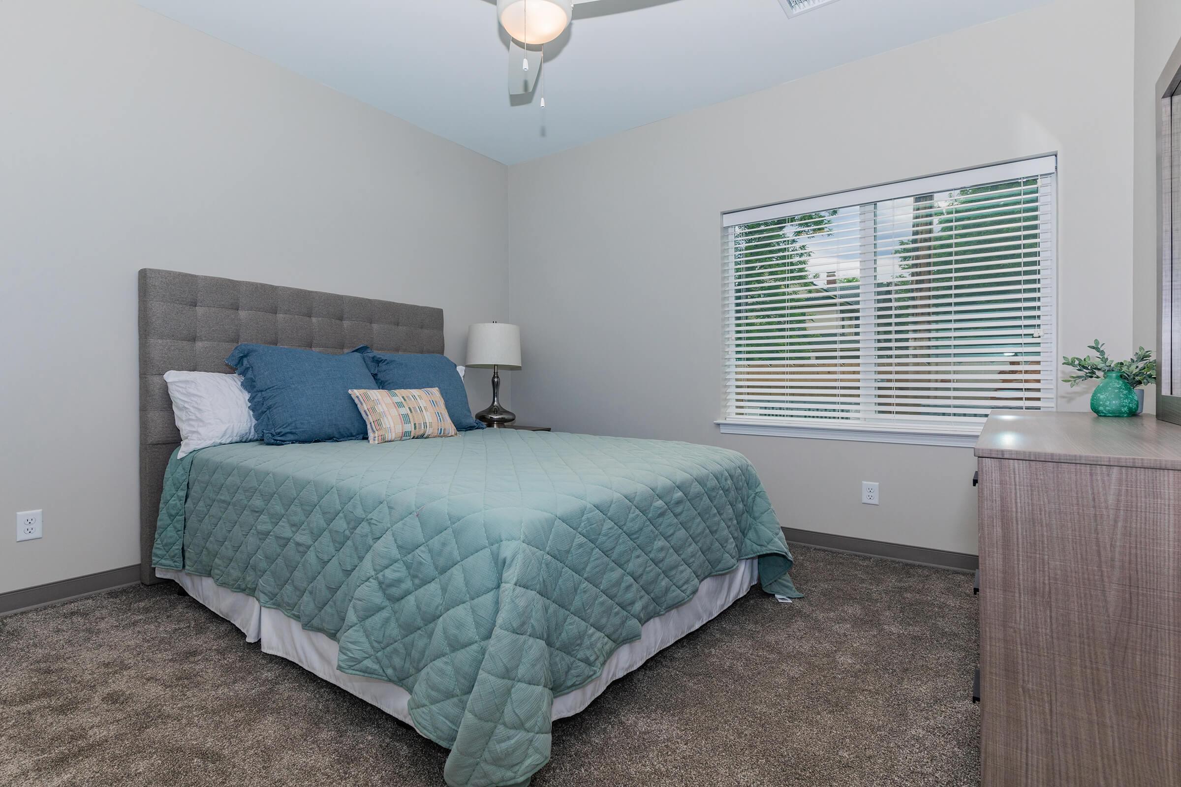 A cozy bedroom featuring a large bed with a teal quilt and decorative pillows, a bedside lamp on a small table, and a window with blinds allowing natural light. The walls are light-colored, and the floor is covered with beige carpet. A dresser is visible on the right side of the image.