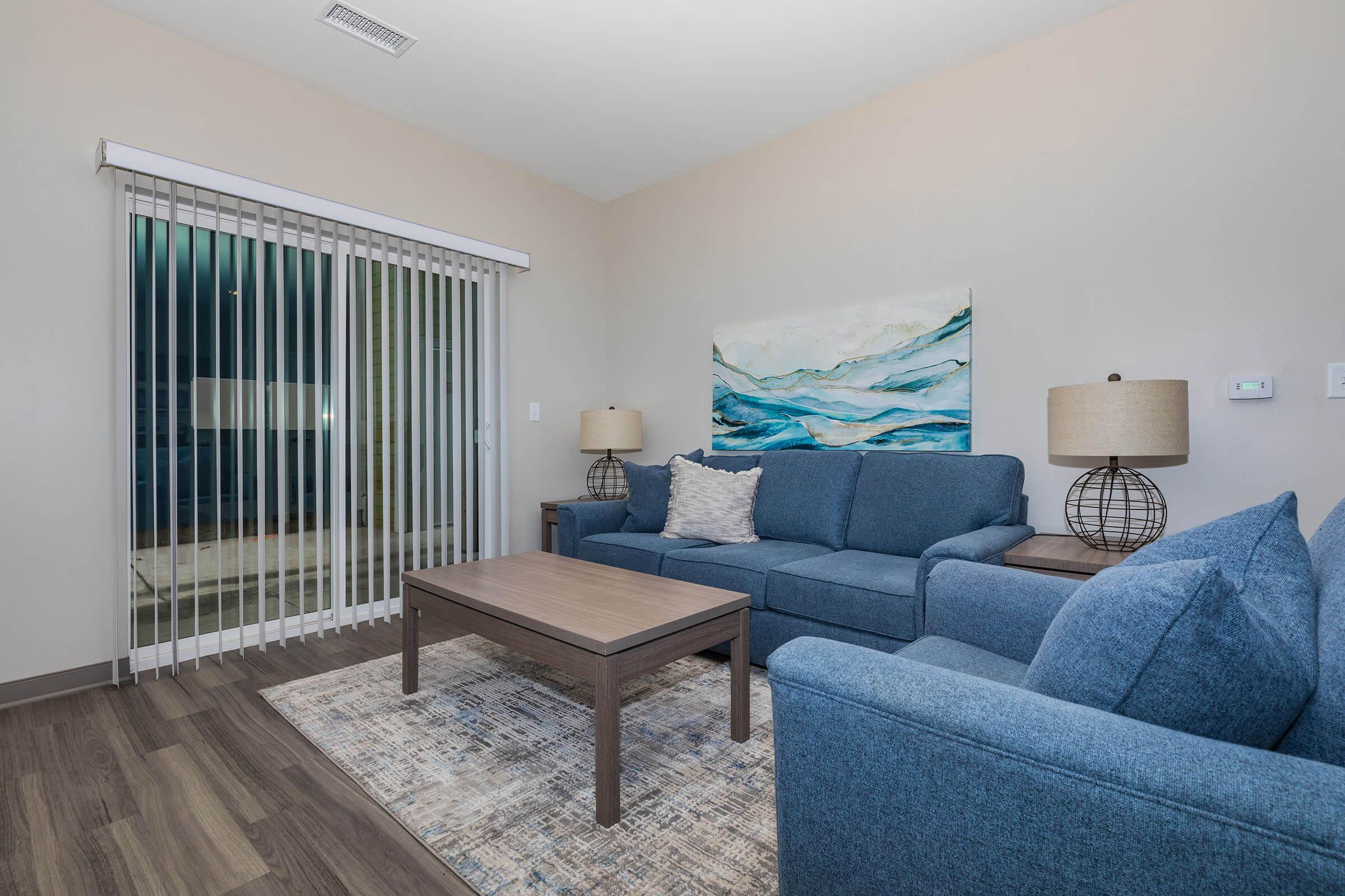 A modern living room featuring a blue sectional couch and matching armchair. A wooden coffee table sits on a patterned area rug. Two lamps with wire frames are on either side of the couch, and a large abstract painting in blue and white hangs on the wall. A glass door with vertical blinds leads to an outdoor area.