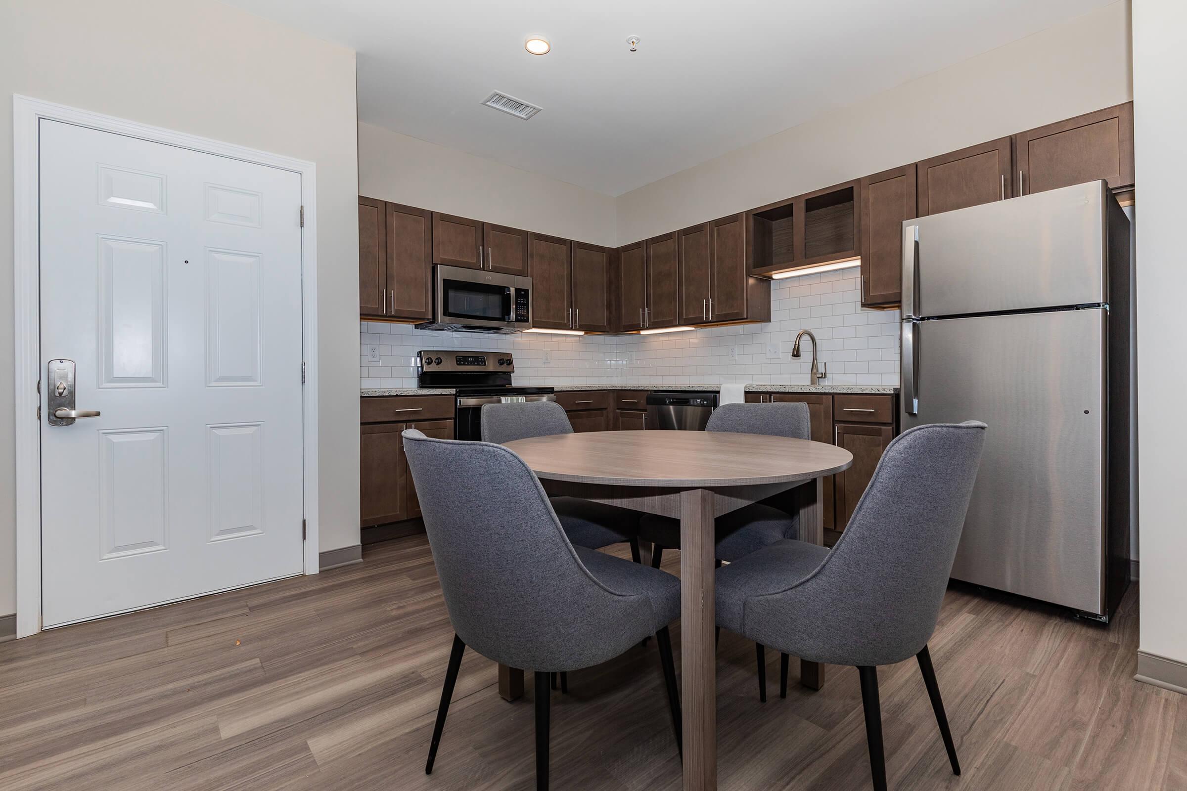 Modern kitchen with dark wood cabinets, stainless steel appliances, and white tile backsplash. A round wooden dining table with four gray upholstered chairs is in the center. A white door is visible on the left, leading to an entrance. The overall decor is contemporary and stylish.