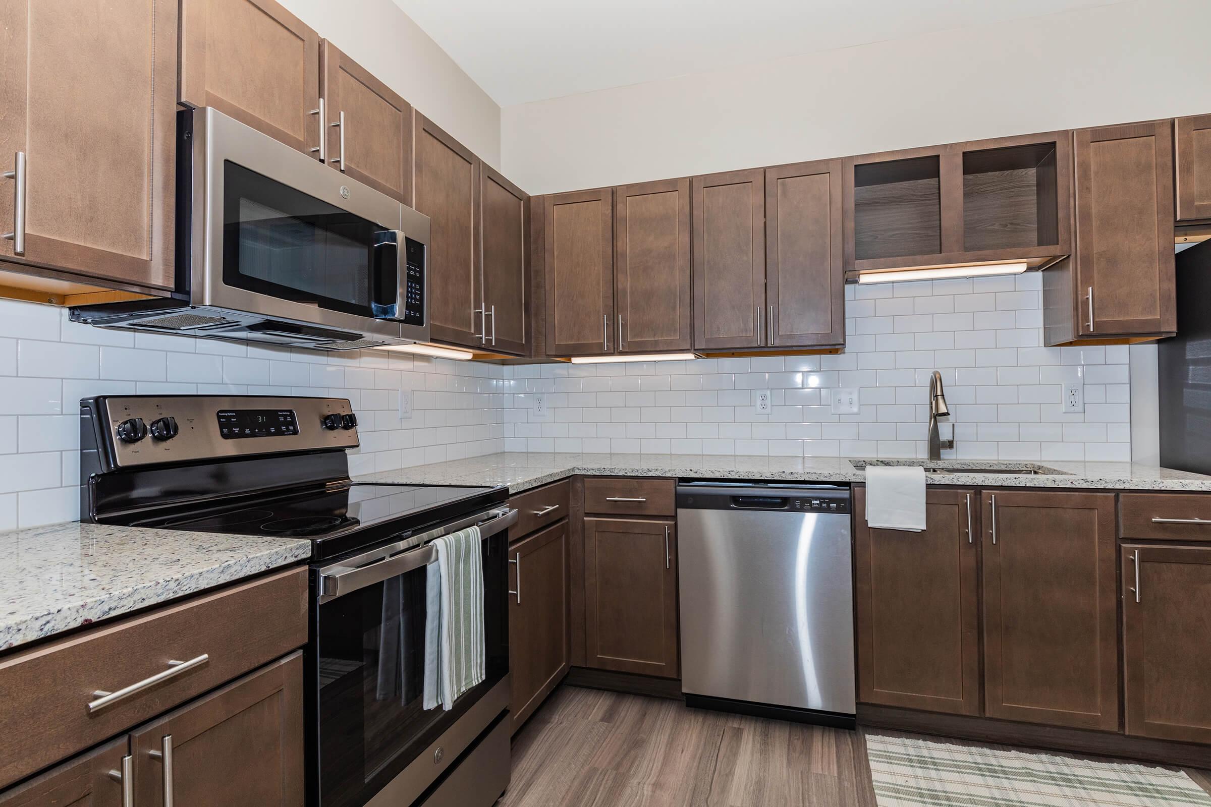 Contemporary kitchen featuring dark wood cabinets, stainless steel appliances including a microwave, oven, and dishwasher, and granite countertops. The backsplash is white subway tile, and there's a sink with a modern faucet. The floor is light-colored wood, creating a warm and inviting atmosphere.