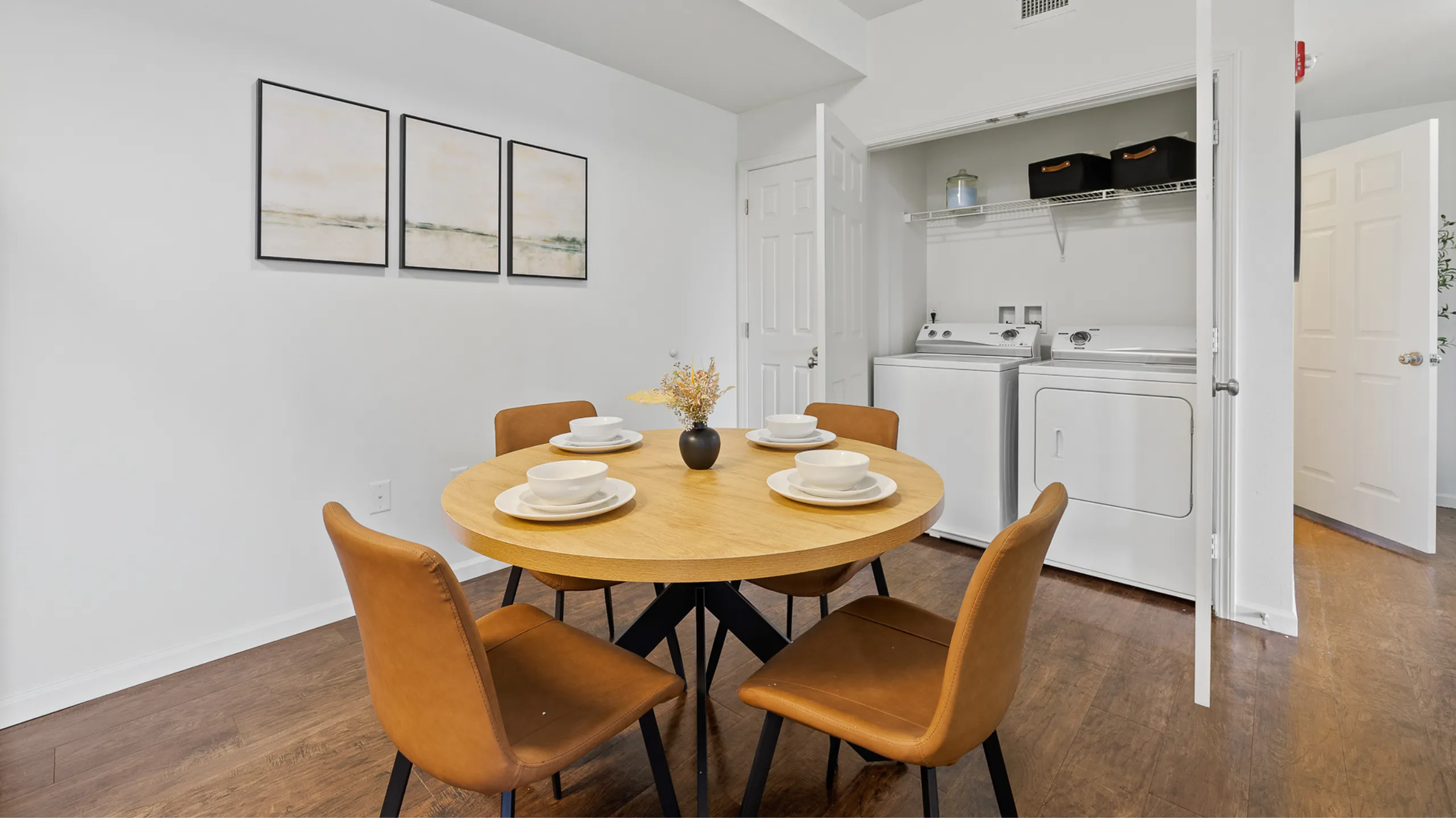 Chic dining area with a round wooden table set for four, accompanied by modern chairs and a view of an in-unit laundry setup, perfect for convenient living.