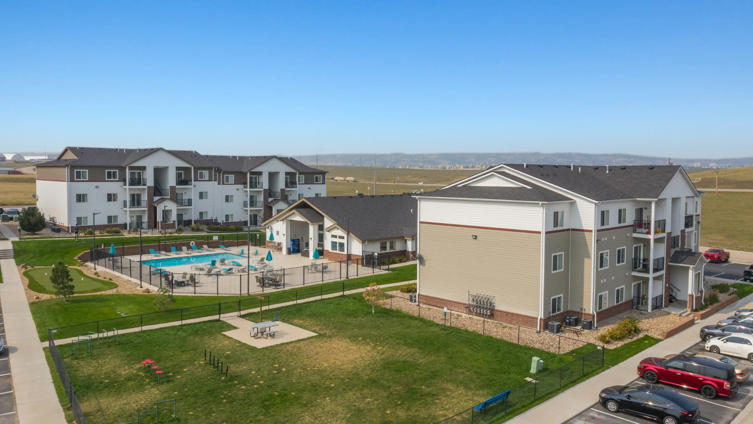 Aerial view of elegant apartment complex featuring modern three-story buildings, a pristine swimming pool, and lush green spaces with scenic mountain backdrop.