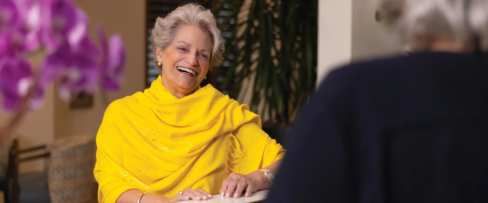 Smiling older woman in a bright yellow outfit chatting warmly across a table, with purple flowers in the foreground.