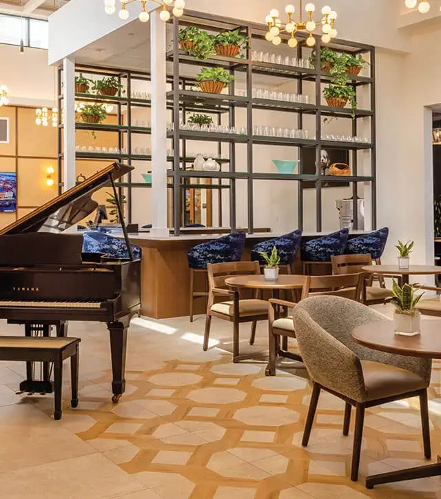 Spacious senior living community lounge featuring a grand piano, modern seating, and tall shelves decorated with greenery.