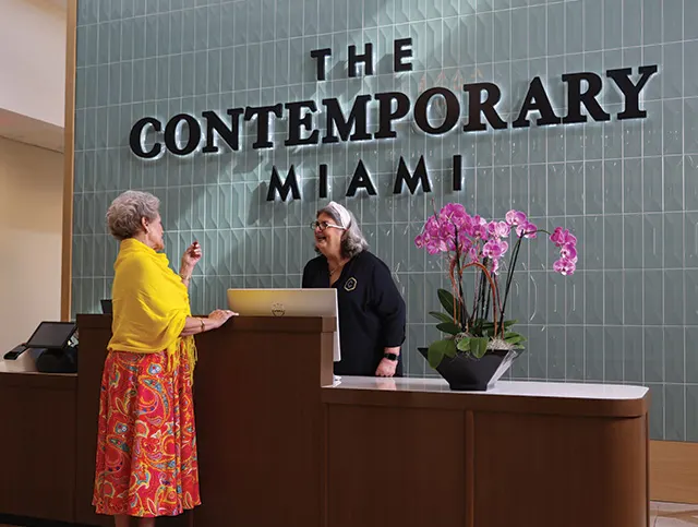 Senior woman speaking with a receptionist at the front desk of The Contemporary Miami, with large signage and orchids in the background.