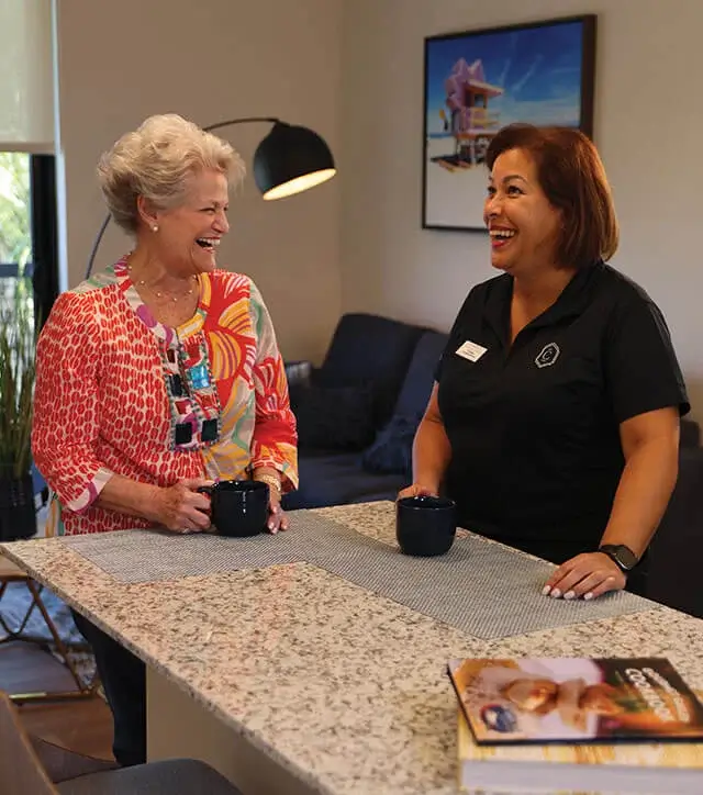 Resident and staff member sharing a laugh over coffee in a modern kitchen area at The Contemporary.