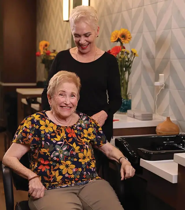 Smiling senior woman seated at a salon chair with a hairstylist standing behind her in the on-site beauty salon.