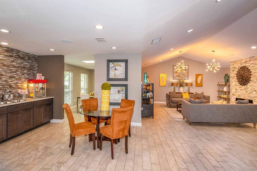 A modern, open-concept living space featuring a dining area with a circular table and orange chairs. To one side, a kitchenette with a decorative backsplash and popcorn machine. The lounge area includes a cozy seating arrangement with gray sofas, stylish decor, and vibrant lighting fixtures, all in a bright, inviting atmosphere.