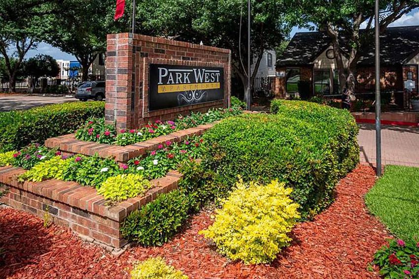 A brick sign for Park West, surrounded by vibrant green shrubs and colorful flower beds. The landscaping features red mulch and a variety of plants, creating an inviting appearance. In the background, trees provide shade, and vehicles are parked nearby.