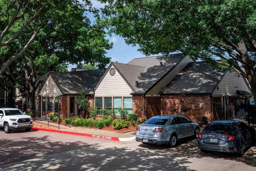 A single-story building with a mix of brick and siding exterior, surrounded by lush greenery and trees. The foreground features a red curb with parked cars, including a white truck and a silver sedan, under a clear blue sky. The overall scene conveys a well-maintained environment.