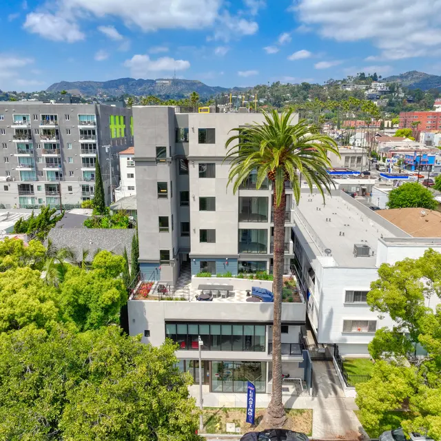 A modern multi-story apartment building surrounded by palm trees and greenery, with a view of the hills in the background under a partly cloudy sky.