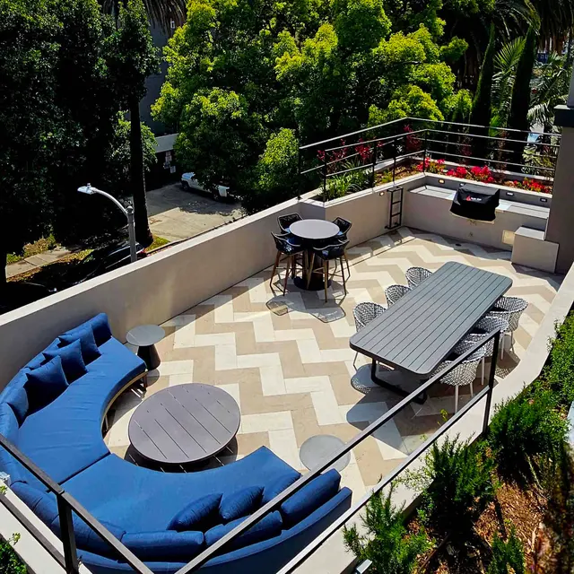 A spacious rooftop lounge area featuring a blue sectional sofa, a round coffee table, a long dining table, and bar stools, surrounded by greenery and large palm trees.