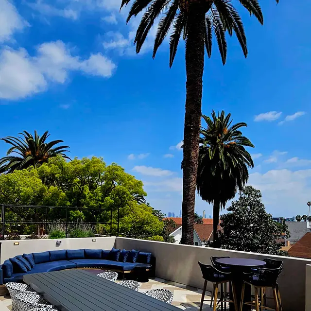 A serene outdoor patio with a geometric patterned floor, featuring stylish lounge furniture, a dark table, and palm trees against a bright blue sky with scattered clouds.