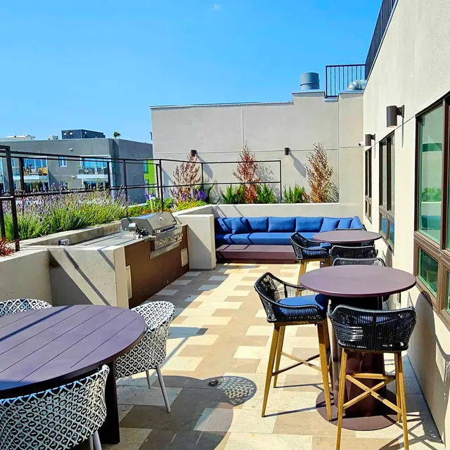 A modern rooftop lounge area with furniture including a blue couch, round tables, and chairs, surrounded by greenery and buildings in the background.