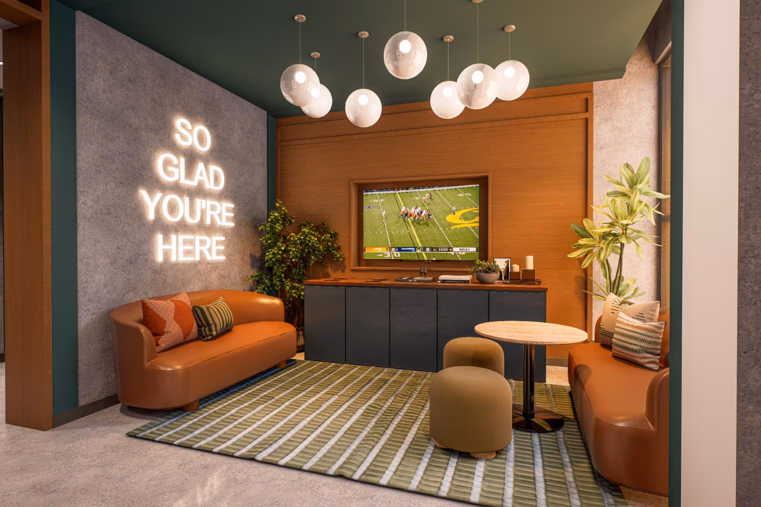 A modern resident common area with a television screen, a neon sign that says "SO GLAD YOU'RE HERE," and comfortable orange seating, set against accent walls with wood paneling and dark teal trim.