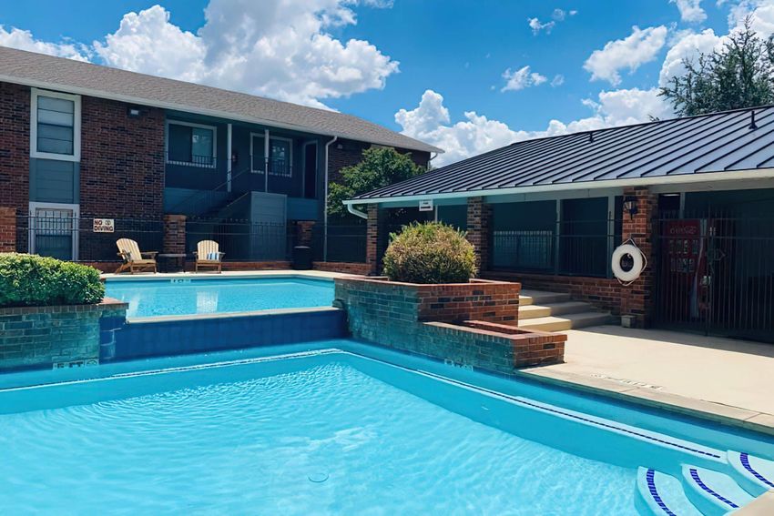 A clear blue swimming pool surrounded by brick and greenery, with a multi-story brick building in the background. The scene features sun loungers near the pool and a shaded area with a roof. White clouds are visible in a bright blue sky.