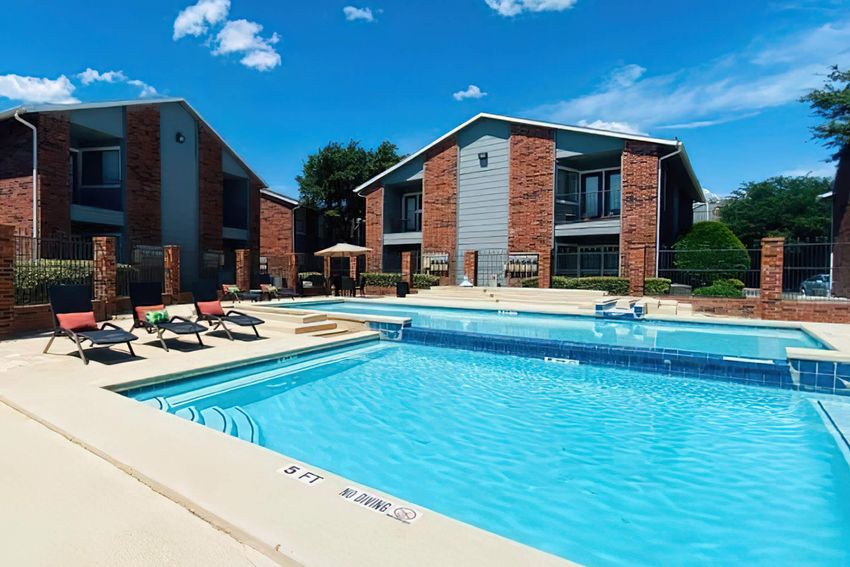 A sunny outdoor swimming pool area surrounded by brick apartment buildings. The pool features a shallow end marked with a "5 FT" sign, and there are lounge chairs positioned nearby. The area is well-lit, with clear blue skies and a few clouds in the background.