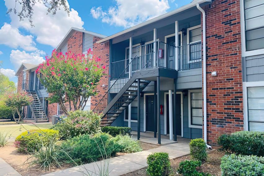 A view of an apartment community featuring two-story buildings with brick exteriors, a set of stairs leading to upper units, landscaped greenery, and flowering shrubs under a blue sky with clouds.